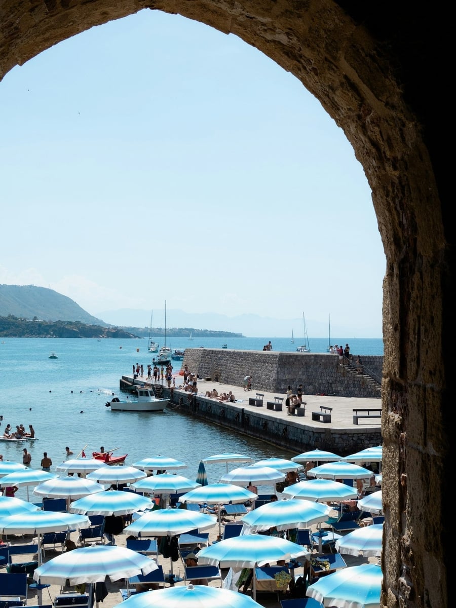 La spiaggia vista dal bastione — Ombrelloni, barche a vela e il mare aperto incorniciati da un arco medievale